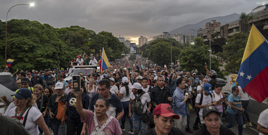 A large crowd of protesters marches through a city street, carrying Venezuelan flags and banners, advocating for democracy and civil rights.
