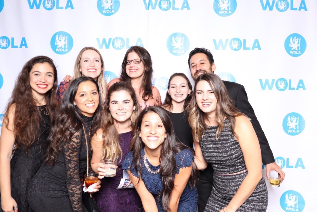 A group of eight individuals poses together in front of a WOLA-branded backdrop, smiling and holding drinks. This photo captures the camaraderie among interns from The Sally Yudelman Internship Program at WOLA.