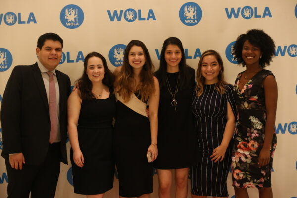 A group of six individuals stands together in front of a WOLA-branded backdrop, all smiling. This photo features interns from The Sally Yudelman Internship Program at WOLA, showcasing their enthusiasm.