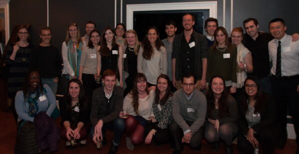 A large group of approximately 30 individuals poses for a photo at a gathering in an indoor setting. The attendees, many of whom are likely participants in The Sally Yudelman Internship Program at WOLA, are smiling and engaging with one another.