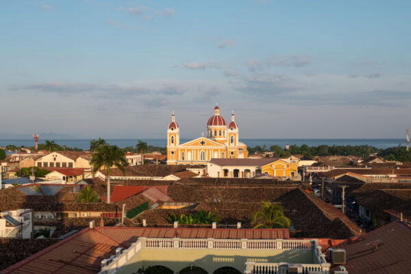 A panoramic view of Granada, Nicaragua, featuring the vibrant yellow Cathedral of Granada with red domes, surrounded by colonial-style rooftops and a distant lake.