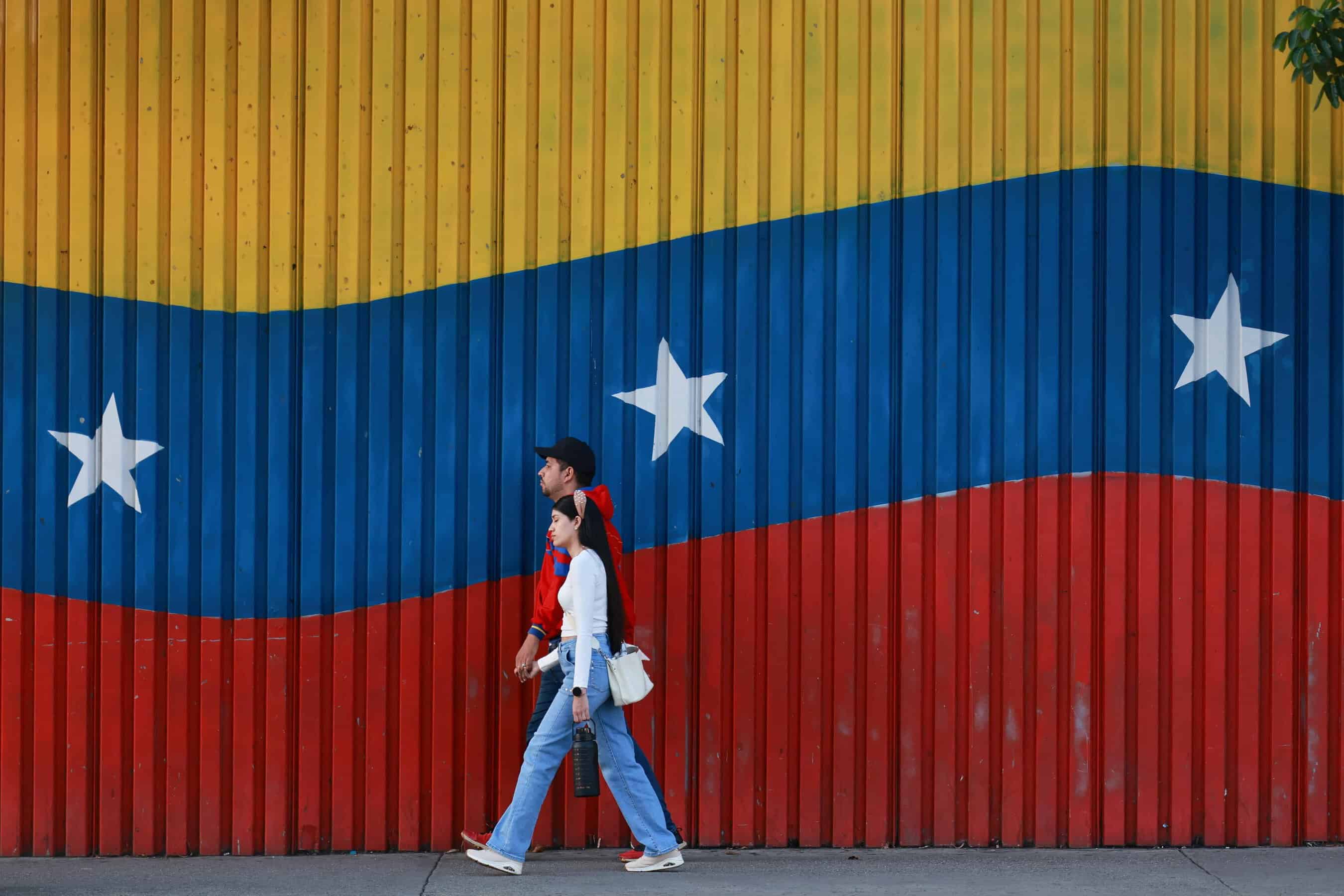 Two people walk past a corrugated metal wall painted with the Venezuelan flag, featuring yellow, blue, and red stripes with white stars.